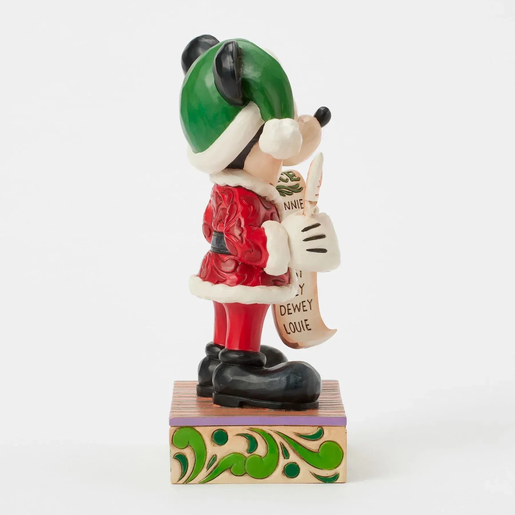 Een Mickey Mouse-figuurtje in kerstmanpak met groene muts, met een lijst in zijn hand, staat op een versierde houten sokkel.