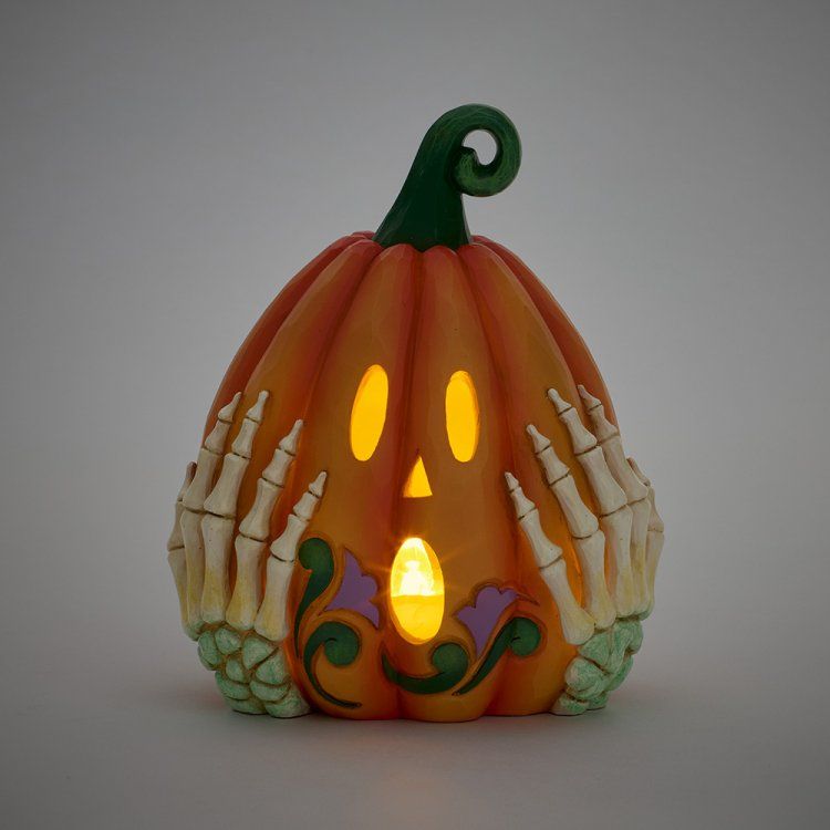 Oranje, verlichte Halloweenpompoen met uitgehouwen gezicht en skeletachtige handen, op een grijze ondergrond.