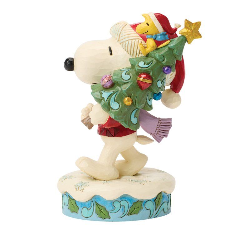 Snoopy met Woodstock op zijn hoofd, die een versierde kerstboom draagt. Ze staan ​​op een besneeuwde ondergrond.