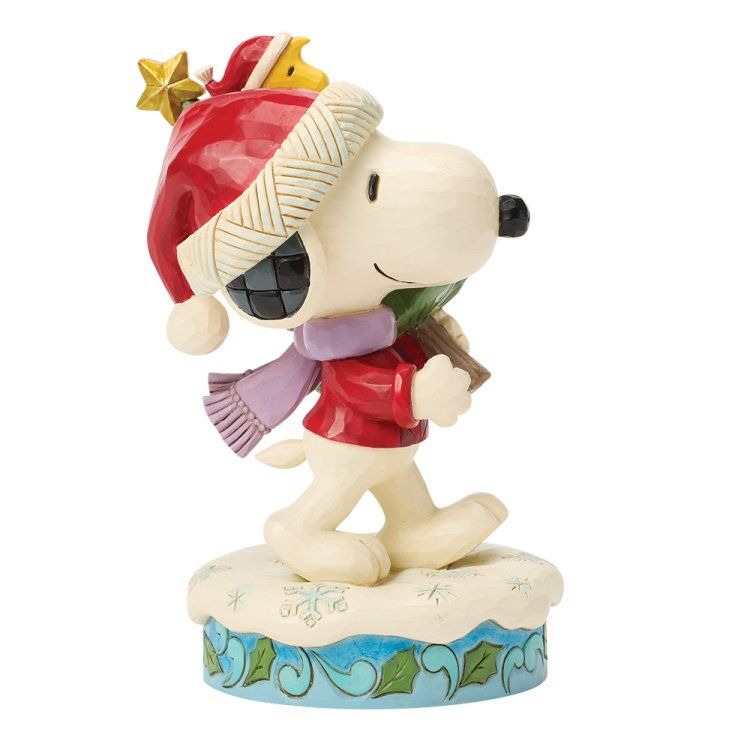 Snoopy in winterkleding draagt ​​een kerstboom, een rode muts met een ster en een sjaal, en loopt over een besneeuwde ondergrond.