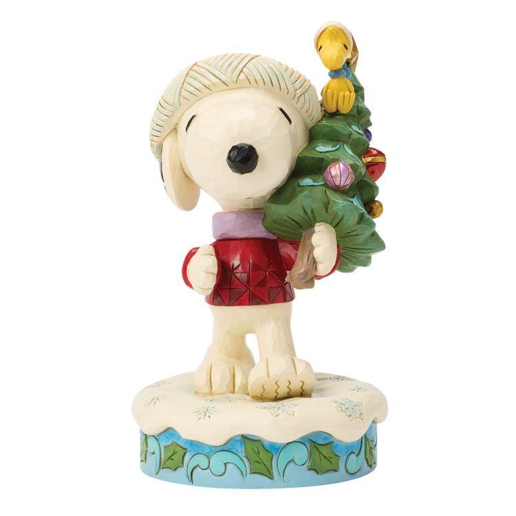 Snoopy, in een rode trui, draagt ​​een kerstboom met Woodstock bovenop, die op een met sneeuw bedekte voet staat.