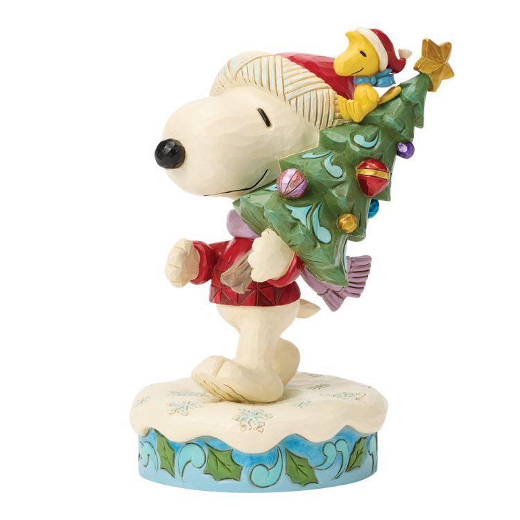 Snoopy, in een rode trui, draagt ​​een kerstboom met Woodstock bovenop, die op een met sneeuw bedekte voet staat.
