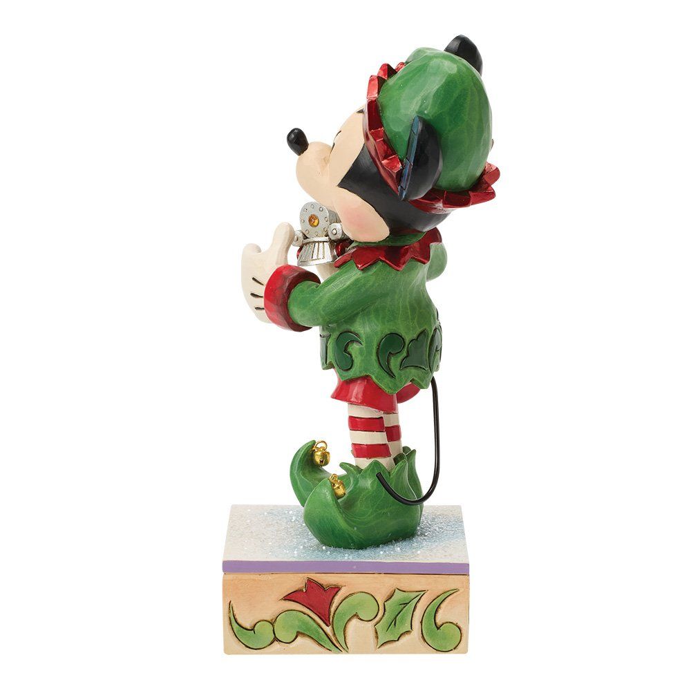 Een Mickey Mouse-elfje met een speelgoedtrein in zijn handen, gekleed in groen en rood, lachend op een voetstuk.