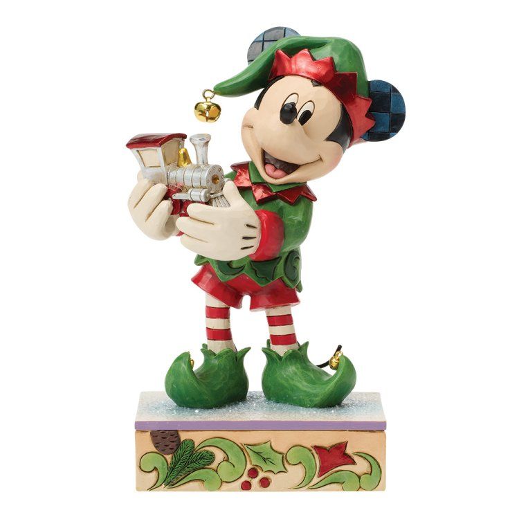 Een Mickey Mouse-elfje met een speelgoedtrein in zijn handen, gekleed in groen en rood, lachend op een voetstuk.