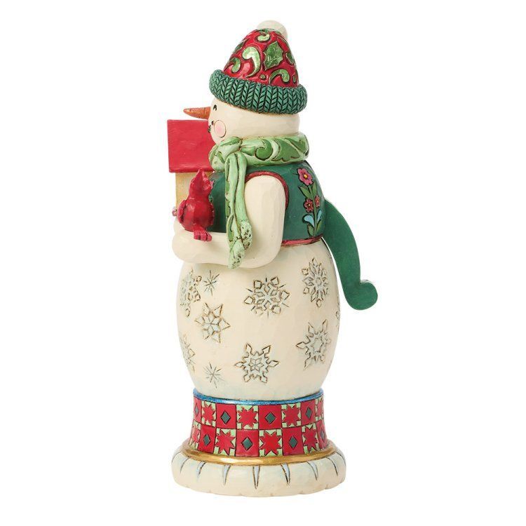 Sneeuwpopfiguurtje met een rood cadeautje en een kardinaal. Hij draagt ​​een groen vest, een groene muts en een groene sjaal en staat op een sokkel met een patroon.
