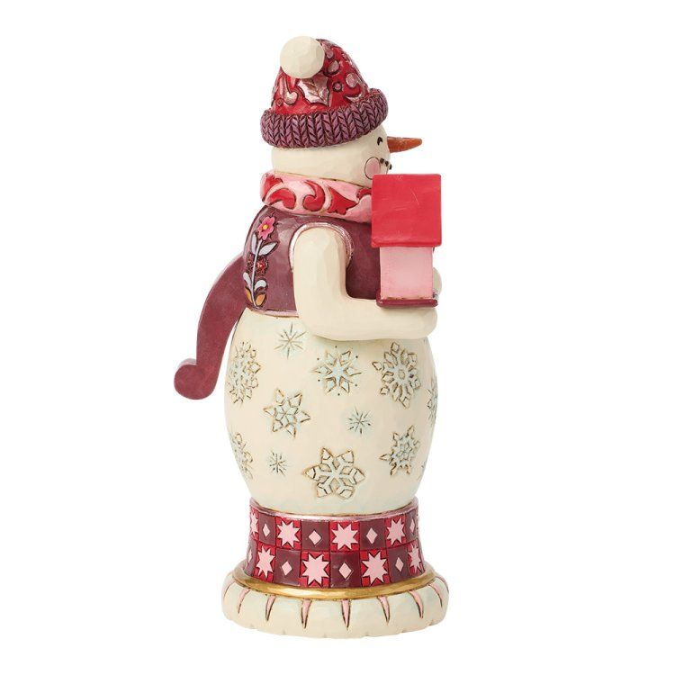Een sneeuwpopfiguur met een rode muts, sjaal en een cadeautje, staand op een versierde sokkel.