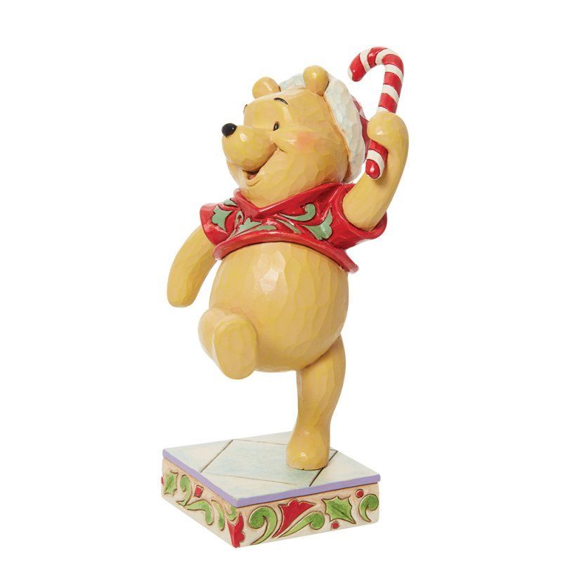 Een Winnie de Pooh-figuurtje in een rood shirt en kerstmuts, met een zuurstok in zijn hand, dansend op een versierde sokkel.