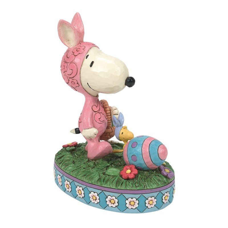 Snoopy in een roze konijnenpak, met een mandje in zijn hand, naast een paasei op een grasveldje.