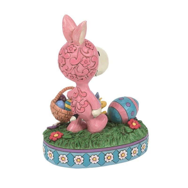 Roze Snoopy als paashaas, met een mandje in zijn hand, staand naast een beschilderd ei op een grasveld.