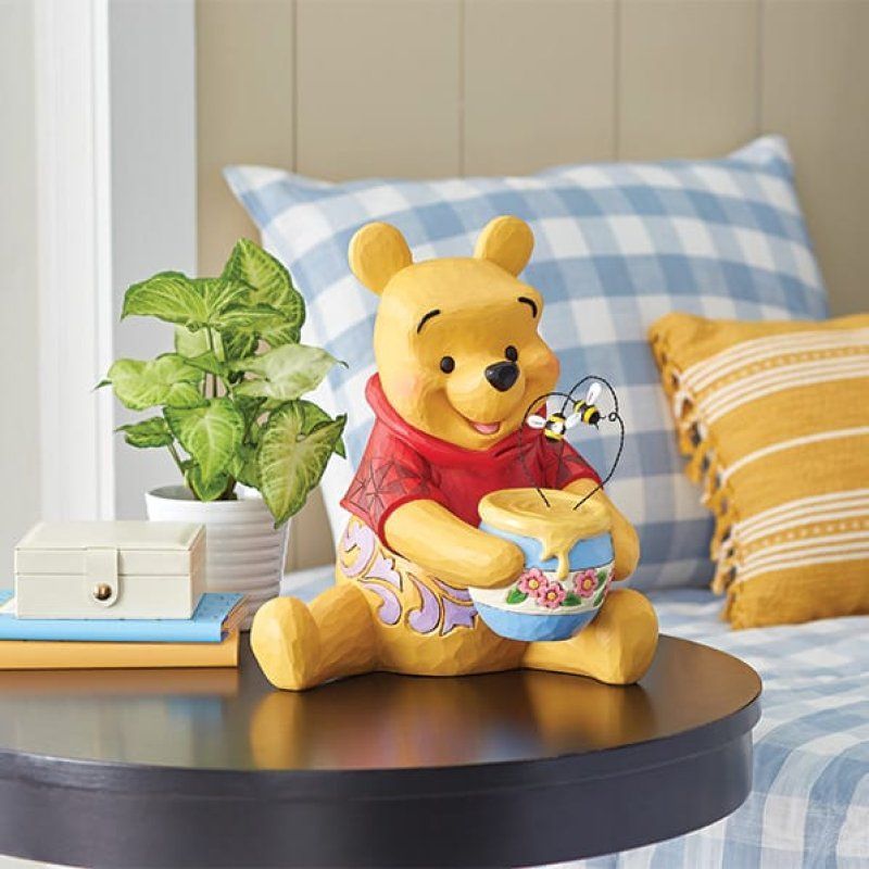Een Winnie de Pooh-beeldje met een honingpot, zittend op een tafel naast een plant, een blauw geruit kussen en een geel kussen.