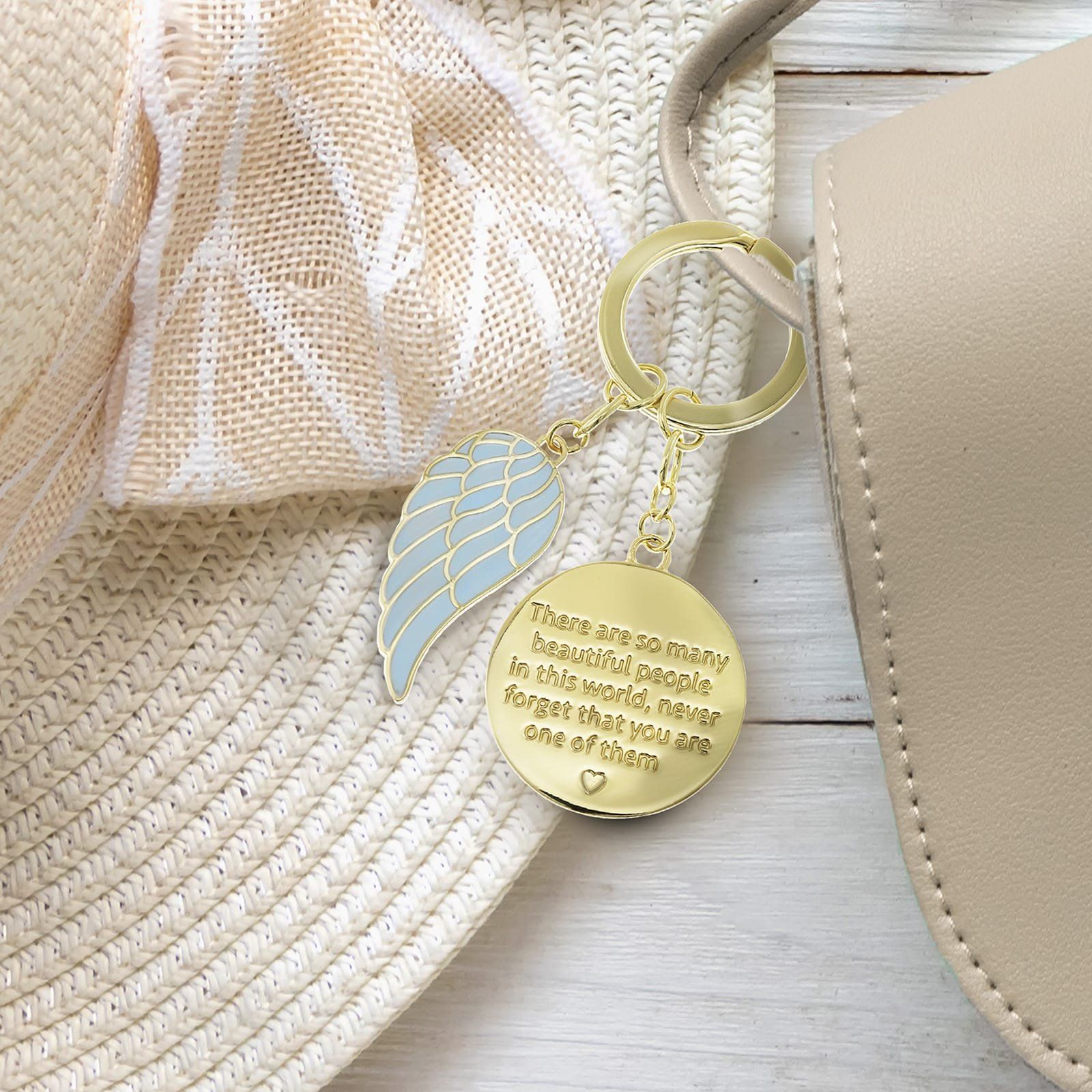 Gouden sleutelhanger met een lichtblauwe vleugelhanger en een ronde hanger met tekst, rustend op een beige hoed en tas.