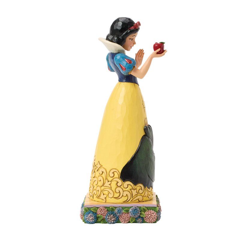 Een standbeeld van Sneeuwwitje met een rode appel in haar hand. Ze draagt een gele jurk, een blauwe top en staat op een sokkel met bloemen.