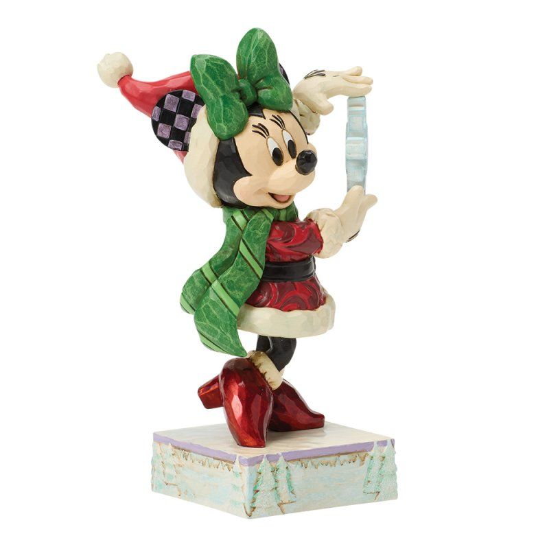 Een kerstfiguurtje van Minnie Mouse met een sneeuwvlokje in haar hand. Groene strik, sjaal, rode jurk en laarzen, staand op een besneeuwde ondergrond.