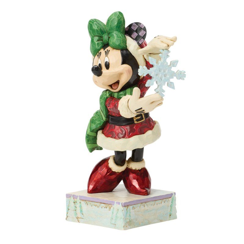 Een kerstfiguurtje van Minnie Mouse met een sneeuwvlok in haar hand, gekleed in een groene sjaal en muts, en een rode jurk, staat op een besneeuwde ondergrond.