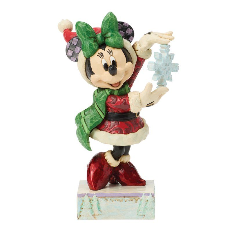 Een kerstfiguurtje van Minnie Mouse met een sneeuwvlok in haar hand, gekleed in een groene sjaal en muts, en een rode jurk, staat op een besneeuwde ondergrond.