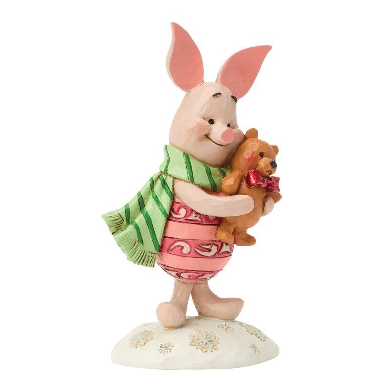 Een beeldje van Knorretje met een teddybeer, gekleed in een gestreepte sjaal en een roze jurk, staand op een met sneeuw bedekte sokkel.