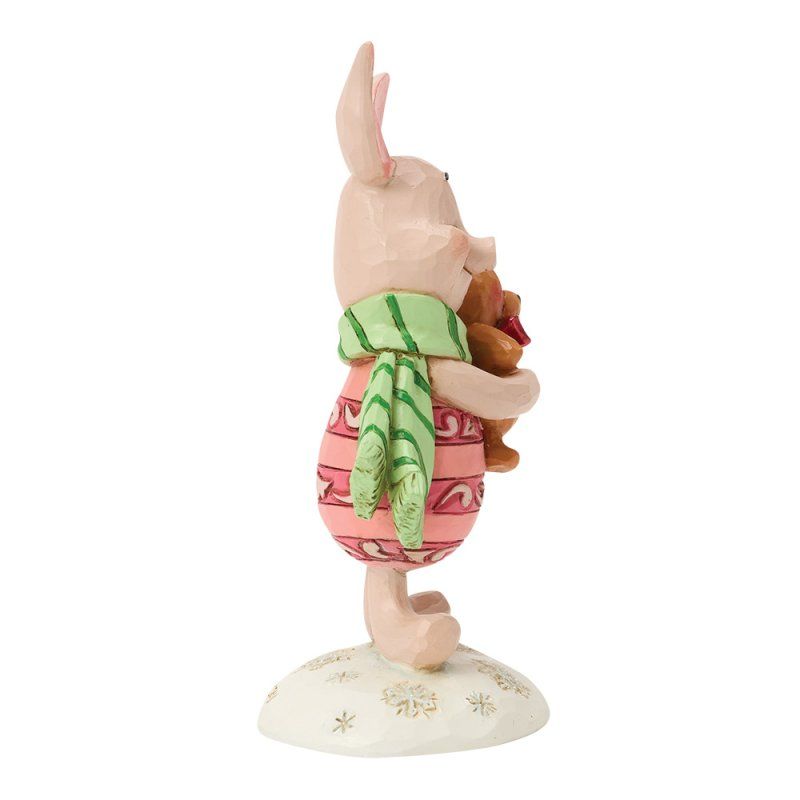 Een beeldje van Knorretje dat een kleine teddybeer vasthoudt, een groene sjaal en een roze trui draagt en op een witte sokkel staat.
