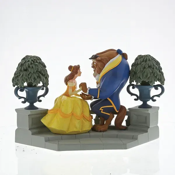 Belle in een gele jurk en het Beest in een blauwe jas, knielend. Ze bevinden zich op een stenen platform met twee potbomen.