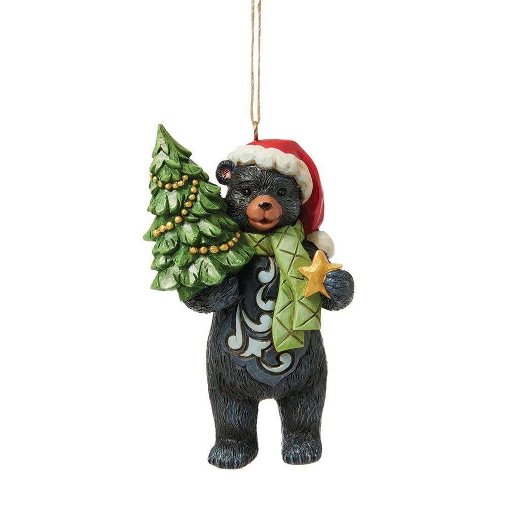 Een zwart beertje met een kerstmuts en -sjaal, dat een klein kerstboompje draagt.