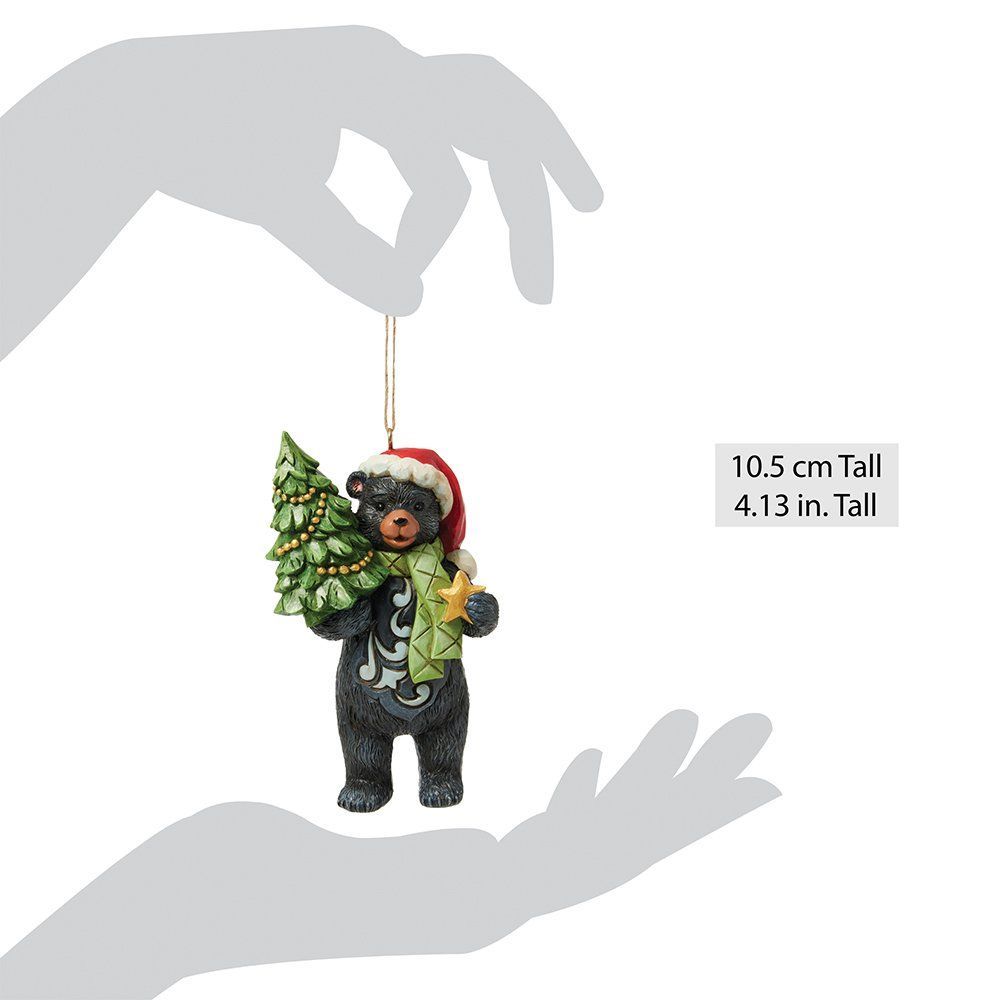 Een zwart beertje met een kerstmuts en -sjaal, dat een klein kerstboompje draagt.
