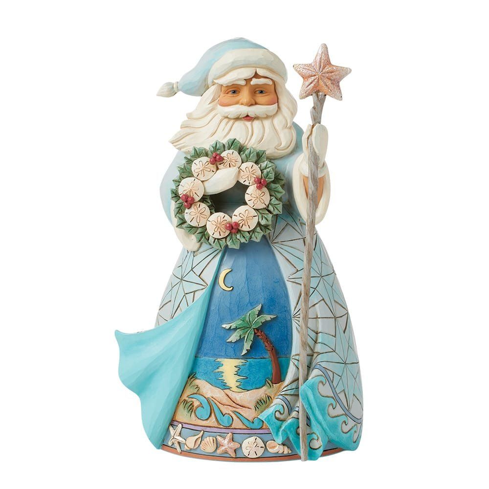Een kerstmanfiguur aan de kust, in een lichtblauwe mantel met een strandtafereel, met een staf en een kerstkrans.