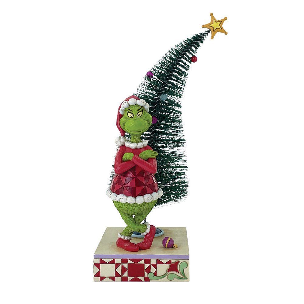 De Grinch in een rode jurk met de armen over elkaar, naast een kerstboom met een ster.