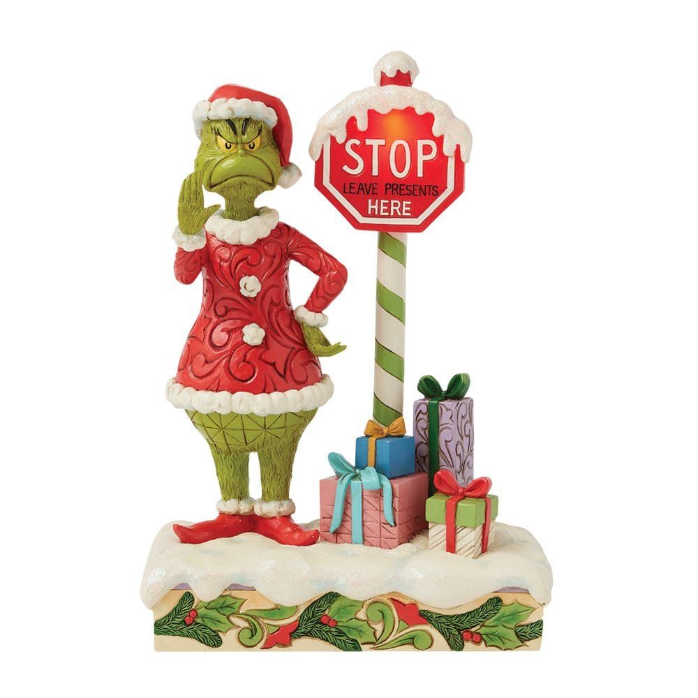 Een Grinch-figuurtje bij een stopbord met cadeautjes, gekleed in een kerstmanpak, die een attent gebaar maakt.
