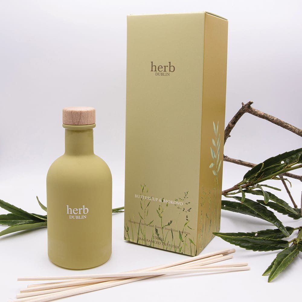 Olijfgroene aromatherapiediffuser met stokjes, naast de doos met kruidenlogo en takjes groen.