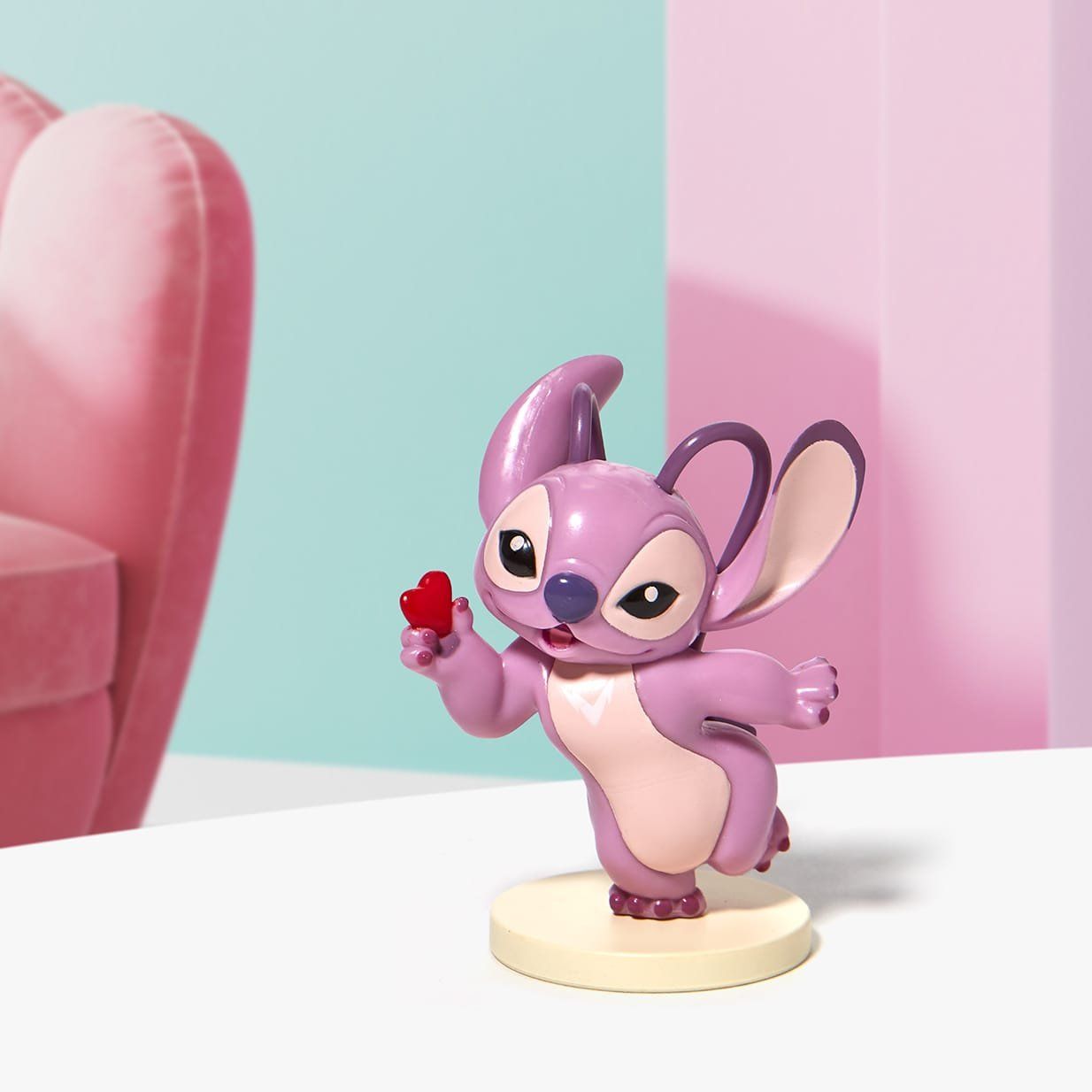 Een lila engeltje dat een rood hartje vasthoudt, staat op een lichtgekleurde schijf. De achtergrond is roze en blauw.