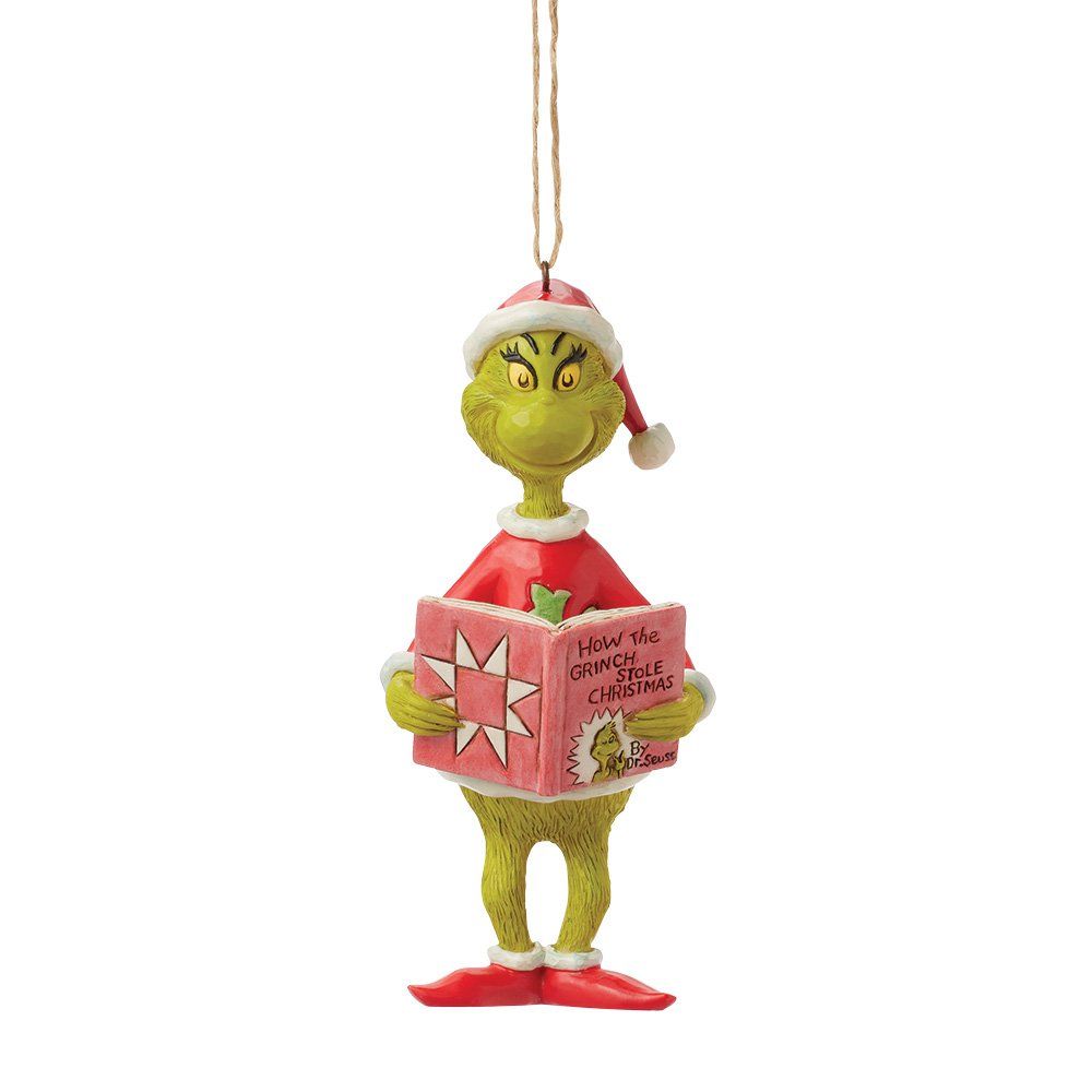 Het kerstornament van de Grinch, die een roze boek vasthoudt, een kerstmuts draagt en een rood pak aan heeft.