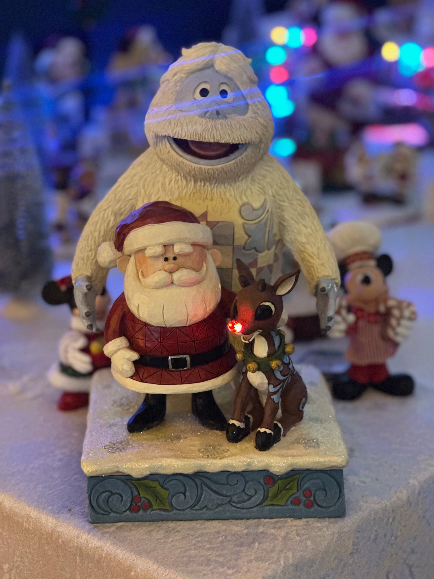 Kerstfiguren: de Kerstman, Rudolf, de Verschrikkelijke Sneeuwman en andere figuren op een besneeuwd platform, met kerstverlichting op de achtergrond.