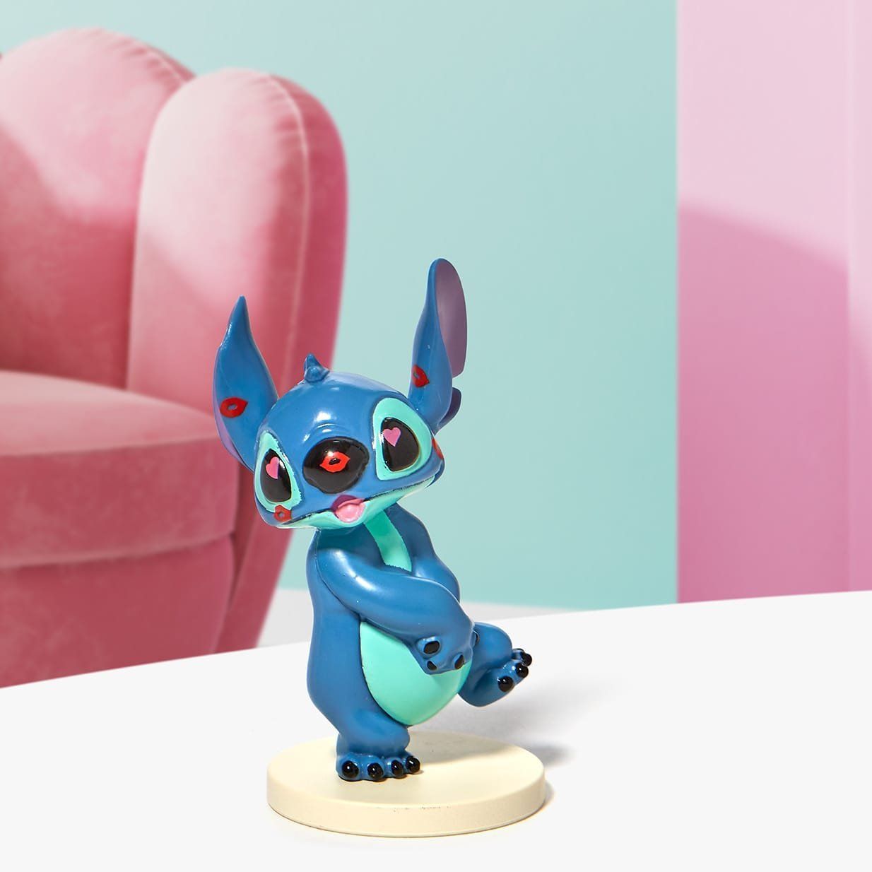 Het beeldje van Blue Stitch staat op een beige sokkel, met een roze stoel en een mintgroene muur op de achtergrond.