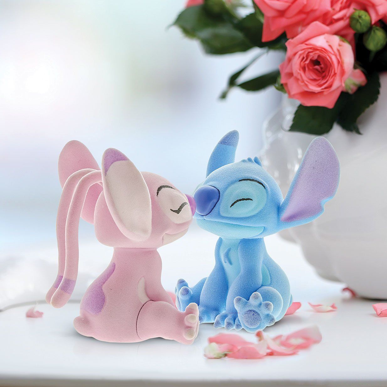 Twee pluche figuren, Angel en Stitch, die elkaar kussen. Rozen op de achtergrond. Roze, blauwe en witte kleuren.