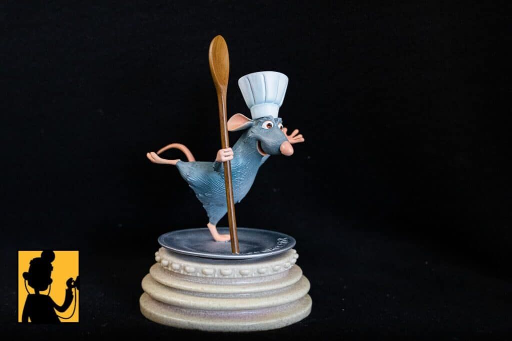 Het Remy-figuurtje uit Ratatouille, met een koksmuts op, balanceert een houten lepel op een bord.