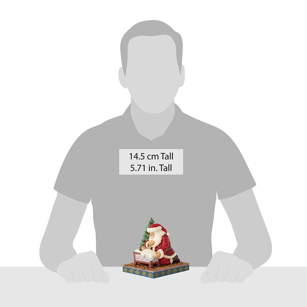 De kerstman knielt en kijkt naar de baby in de wieg, vlakbij een kleine kerstboom. Hij draagt rode en witte kleding.