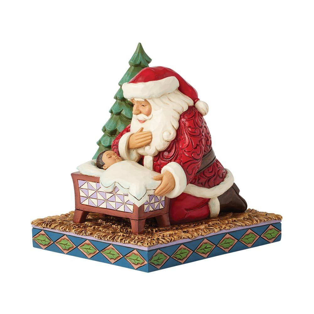 De kerstman knielt en kijkt naar de baby in de wieg, vlakbij een kleine kerstboom. Hij draagt rode en witte kleding.