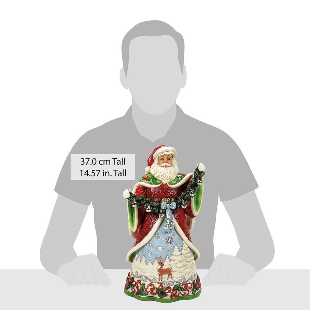 Standbeeld van de Kerstman in een rode mantel, met een kerstkrans in zijn hand. Op de sokkel staan herten en besneeuwde bomen.