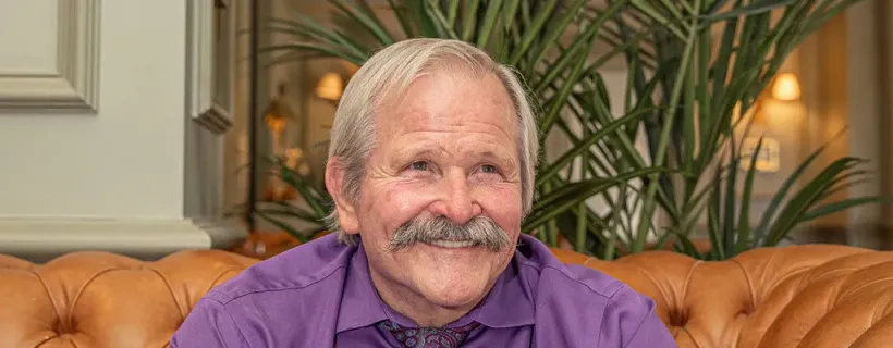 Een man met een witte snor en grijs haar, gekleed in een paars shirt, zit lachend op een oranje leren bank.