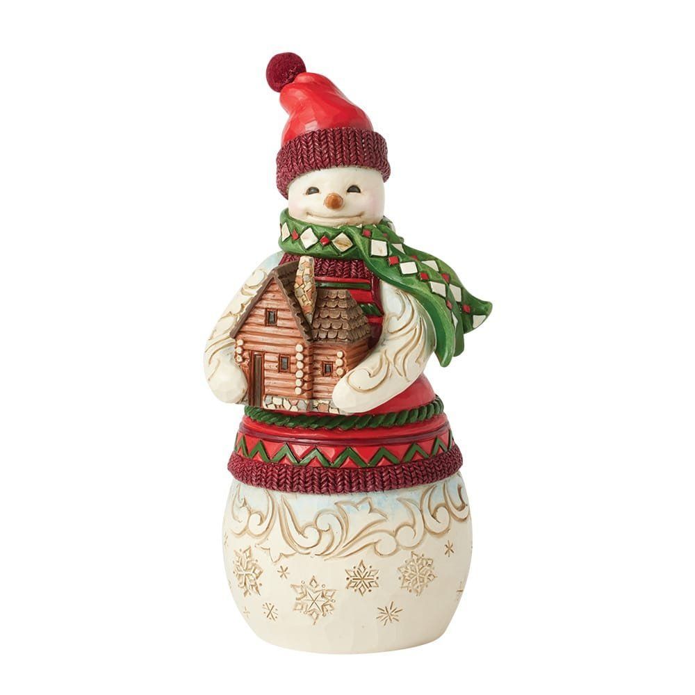 Een sneeuwpopfiguurtje dat een houten hutje vasthoudt. Hij draagt een rode muts en sjaal en lacht.