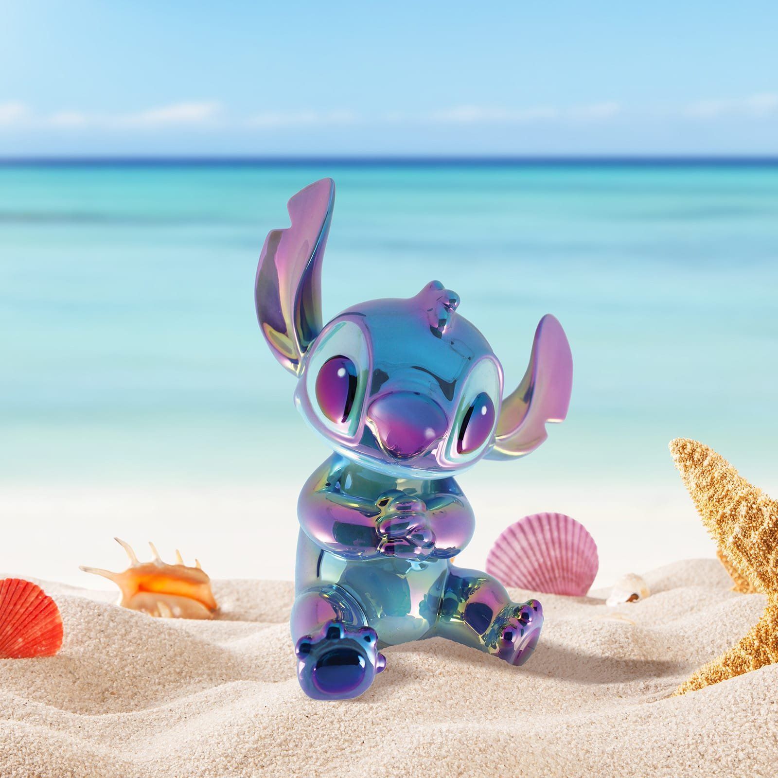 Een iriserend Stitch-figuurtje zit op een zandstrand met schelpen en de oceaan op de achtergrond.