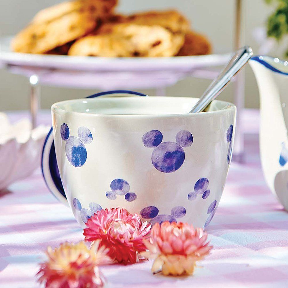Witte mok met blauwe Mickey Mouse-silhouetten, een lepel, roze bloemen en koekjes.