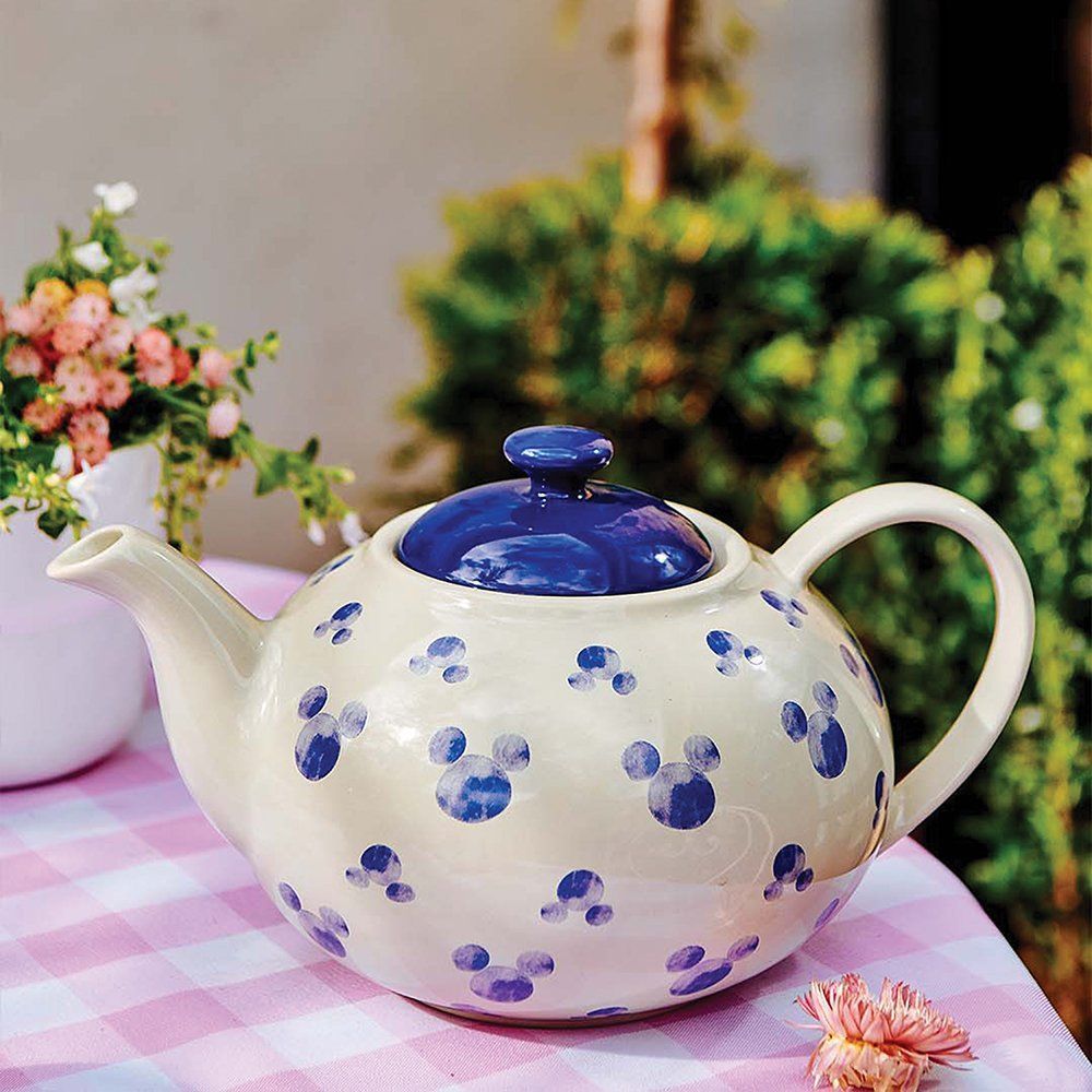 Crèmekleurige theepot met blauwe Mickey Mouse-versieringen, blauw deksel, staat op een roze geruit tafelkleed buiten.