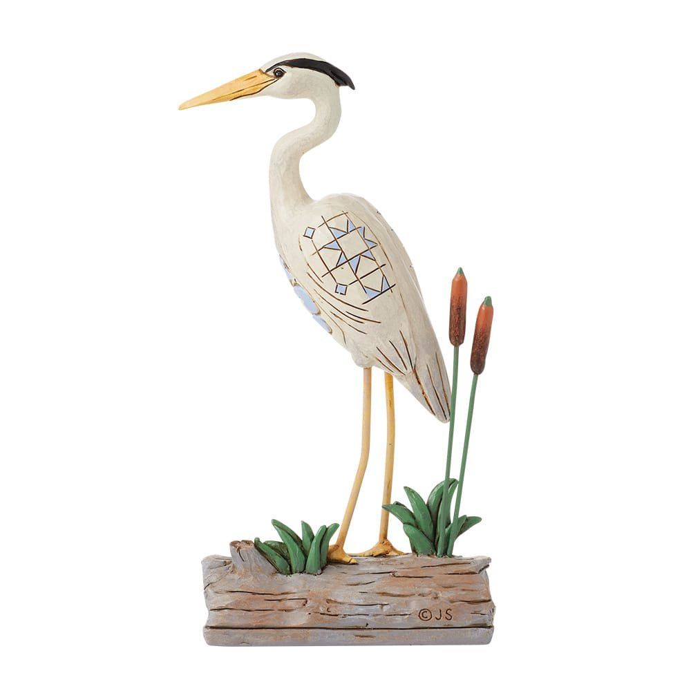 Standbeeld van een reiger die op een boomstam staat, begroeid met riet. De reiger is wit met een gele snavel.