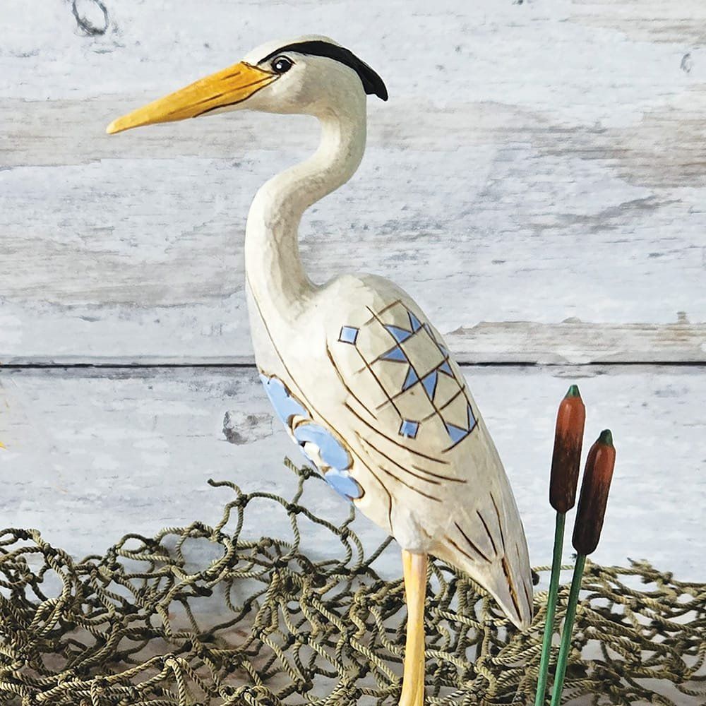 Een uit hout gesneden beeld van een witte reiger met blauwe accenten, staand tussen riet en een visnet.