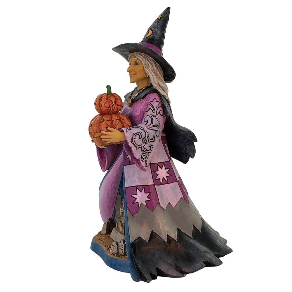 Heksenfiguurtje in een paarse en grijze jurk met een pompoen in haar hand. Ze draagt een zwarte hoed.