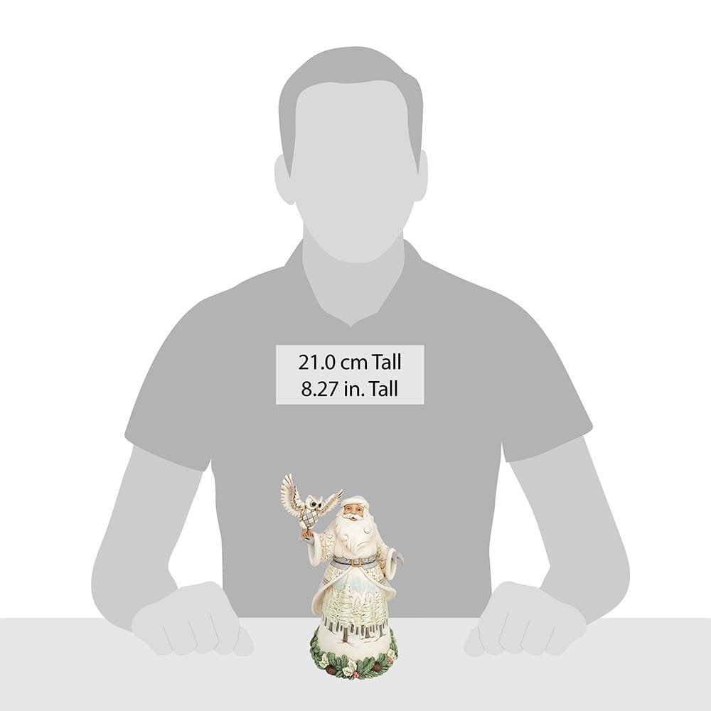 Een kerstmanfiguur met een uil in zijn armen, gekleed in een witte mantel met een boslandschap op de rok, zittend op een bed van groen gebladerte.