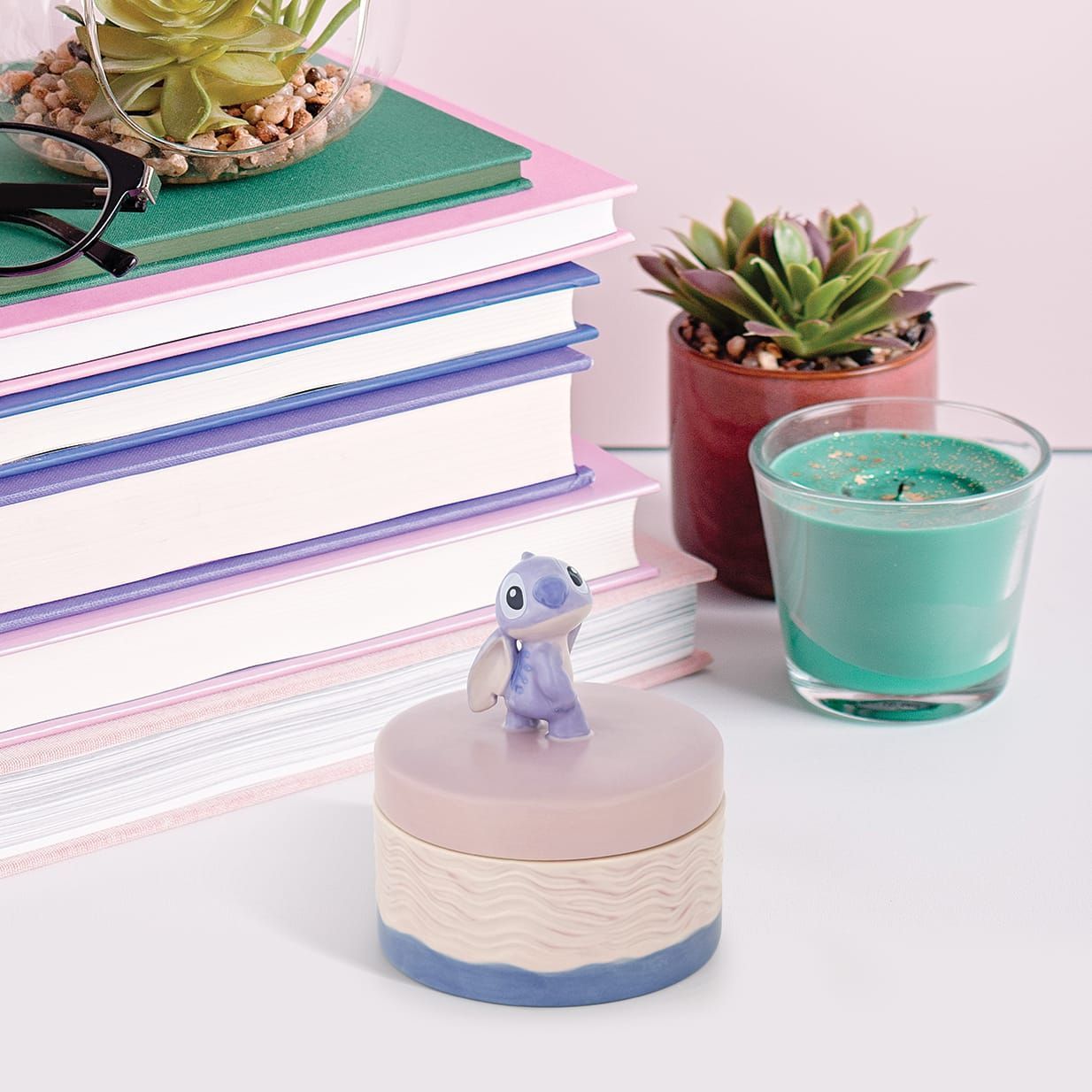 Een Stitch-figuurtje op een klein sieradendoosje, naast een stapel boeken, vetplanten en een turquoise kaars.