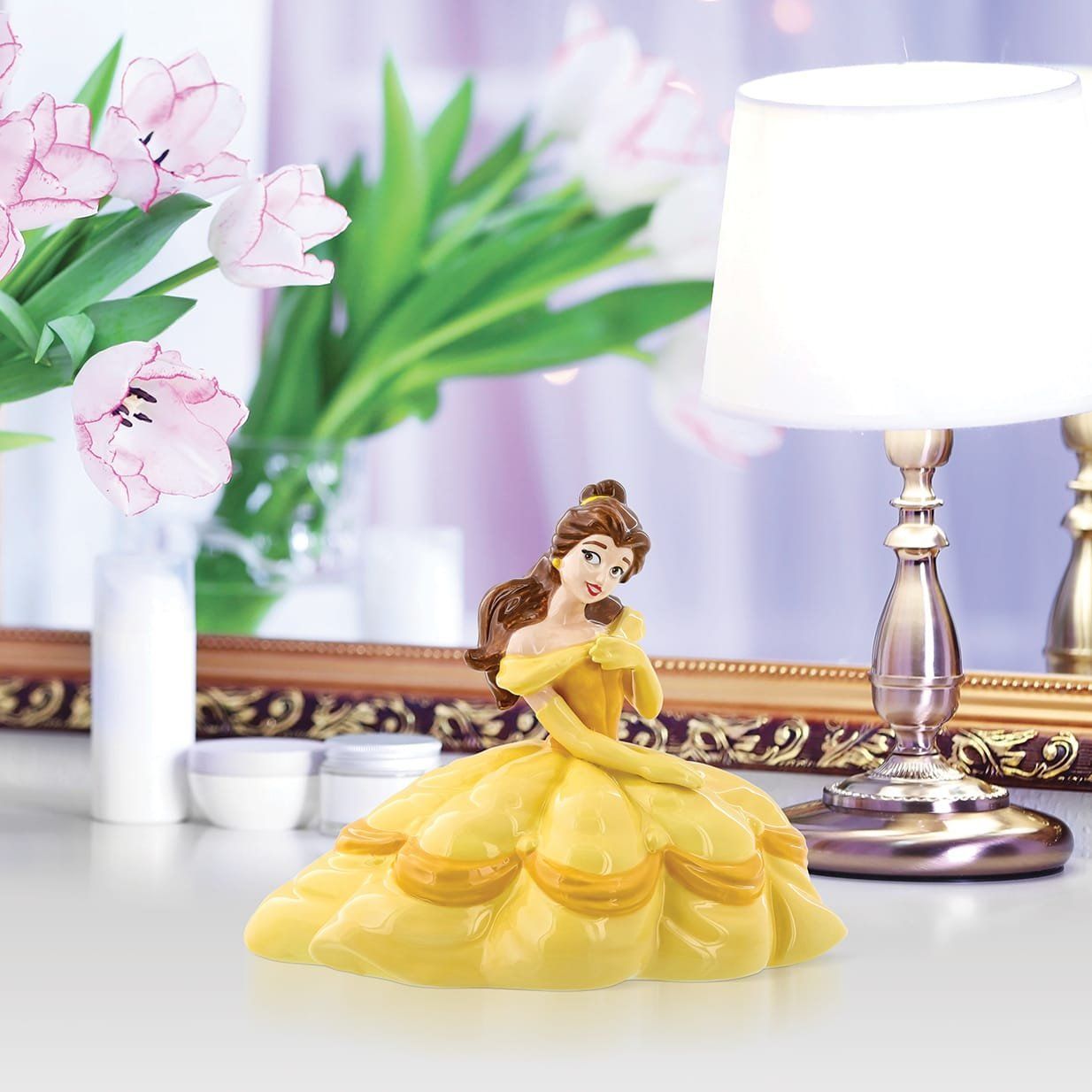 Een beeldje van Belle in een gele jurk, op een tafel, met bloemen en een lamp.