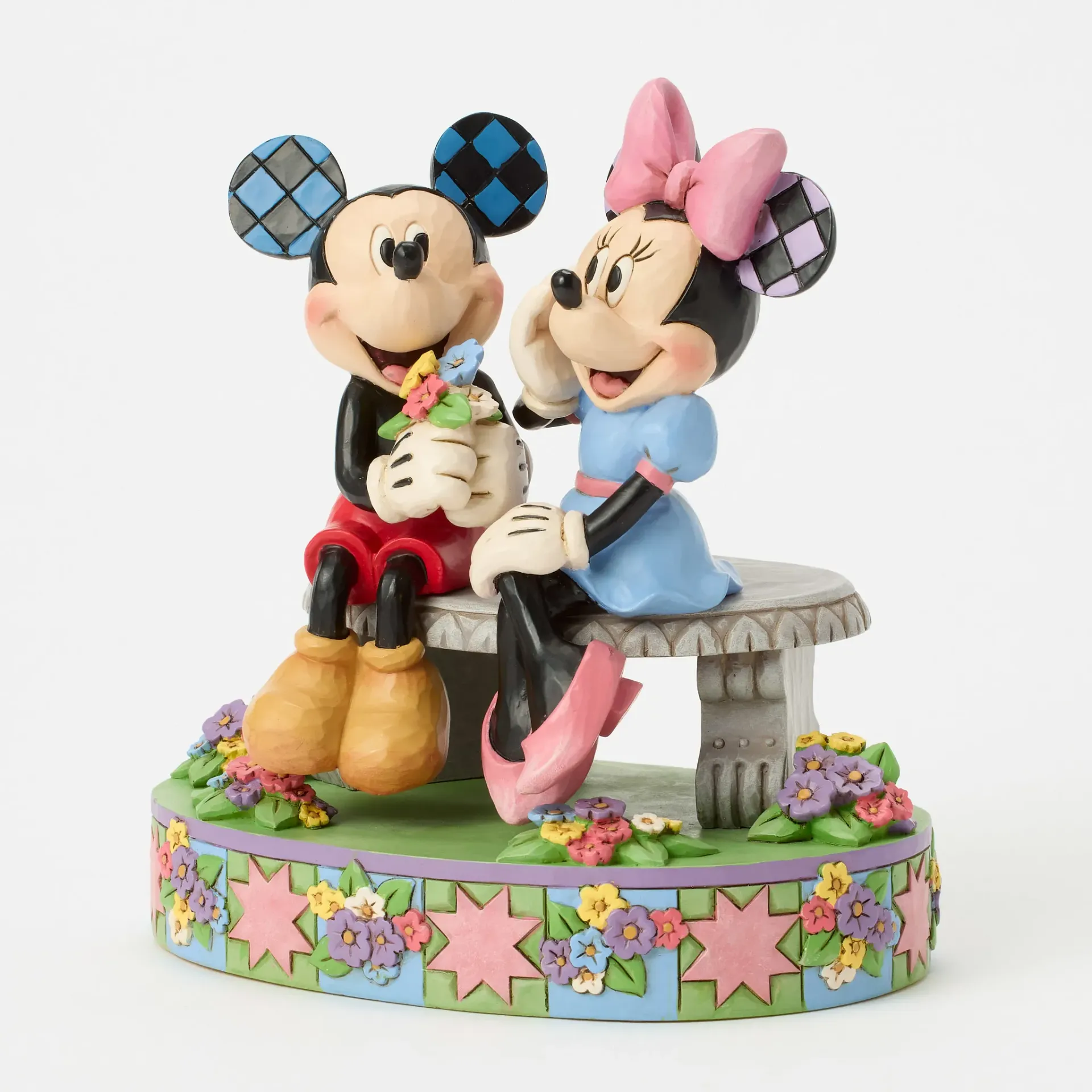 Een beeldje van Mickey Mouse en Minnie Mouse zit op een bankje. Ze zijn omringd door bloemen en hebben oren van patchwork.