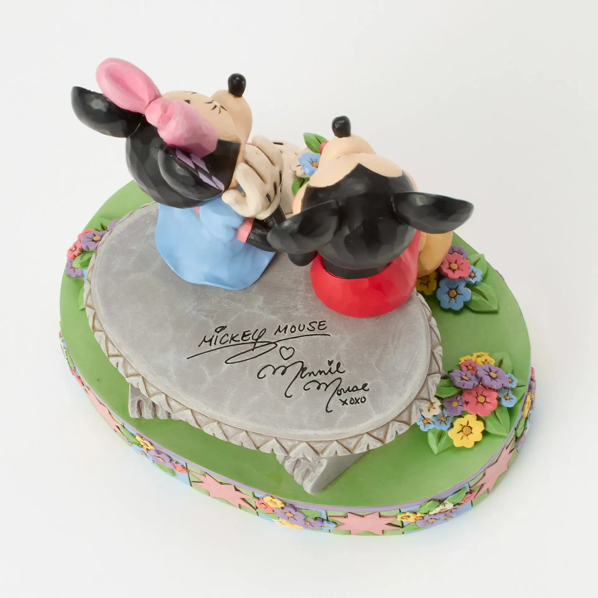 Een beeldje van Mickey Mouse en Minnie Mouse zit op een bankje. Ze zijn omringd door bloemen en hebben oren van patchwork.